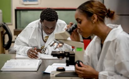 Two students wearing lab coats focused on a microscope in a science lab.