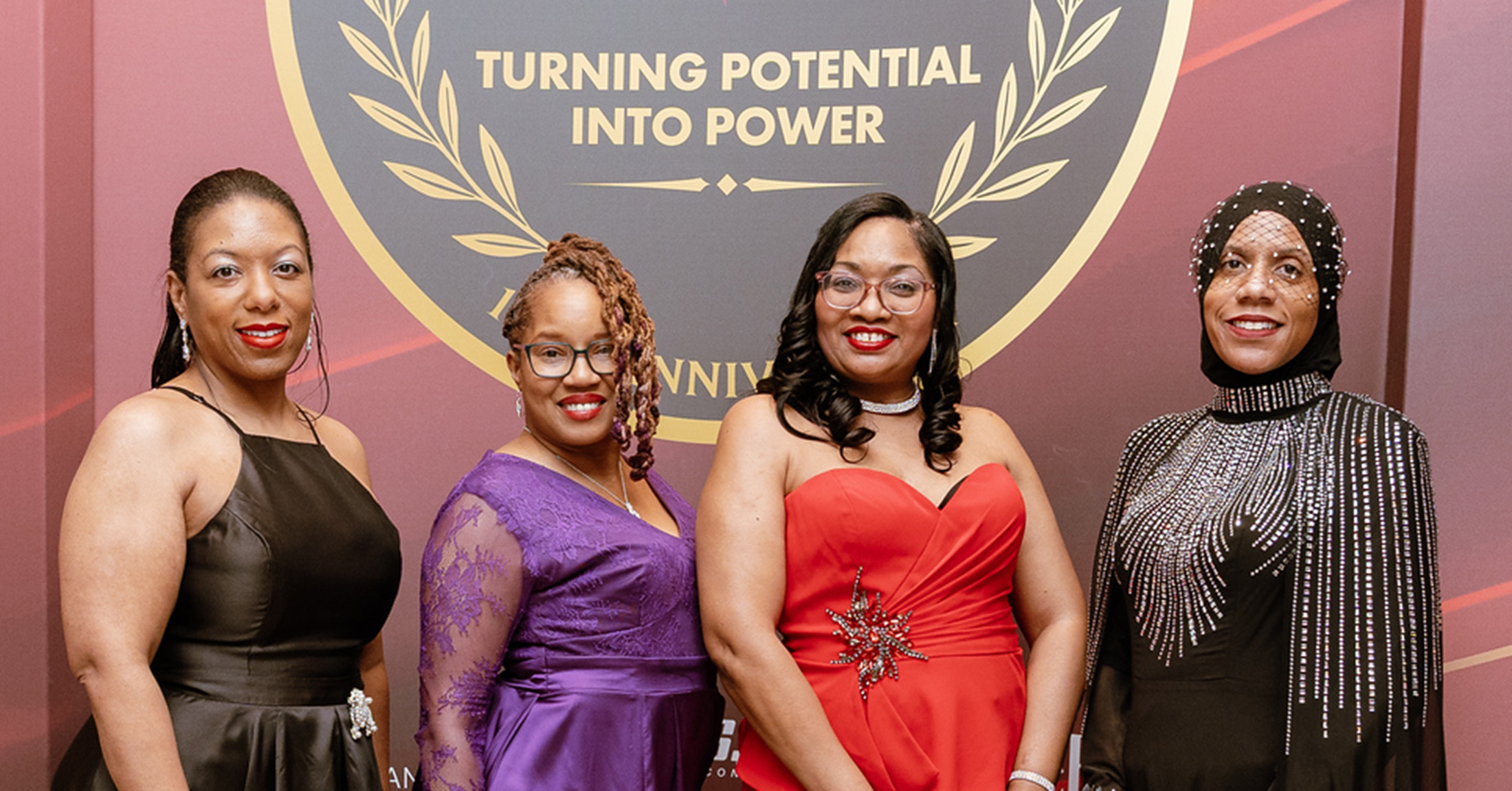 Four women wearing formal gowns smiling in front of a banner that says Turning Potential Into Power.