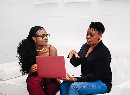 Two african american ladies sitting on couch with laptop having a discussion