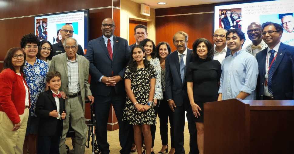 Group photo of diverse individuals standing together, smiling, during a formal event, with a presentation backdrop visible.