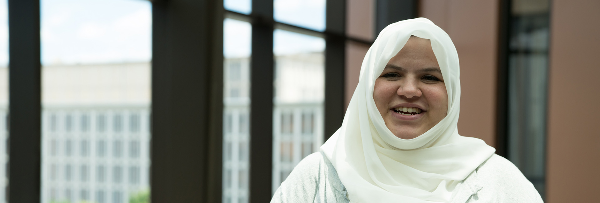 Female student wearing a hijab, standing in front of and window smiling.
