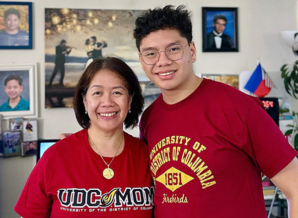 A smiling mother and son pose together, both wearing University of the District of Columbia apparel. The background features family photos and a decorative wall.