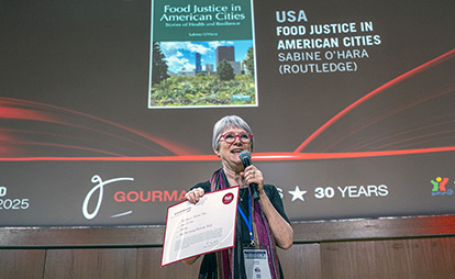 Sabine O'Hara excepting an award and speaking at a microphone on a stage.