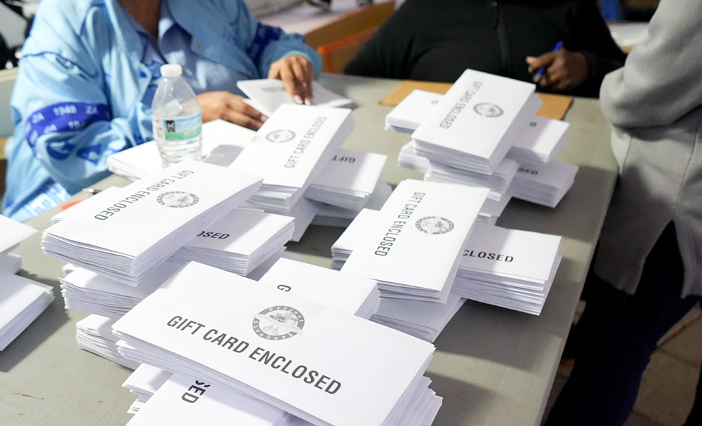 Stacks of envelopes labeled "GIFT CARD ENCLOSED" on a table, with people preparing them in the background.