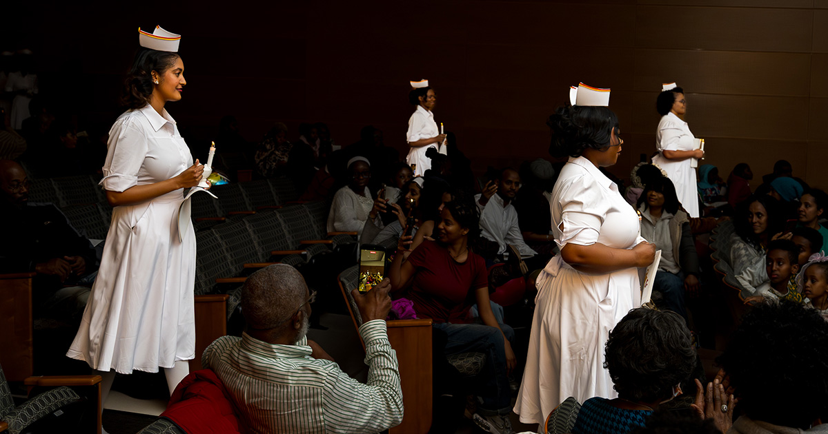 Nursing graduates wearing white nurses uniforms and holding candles in a darkly lit auditorium.