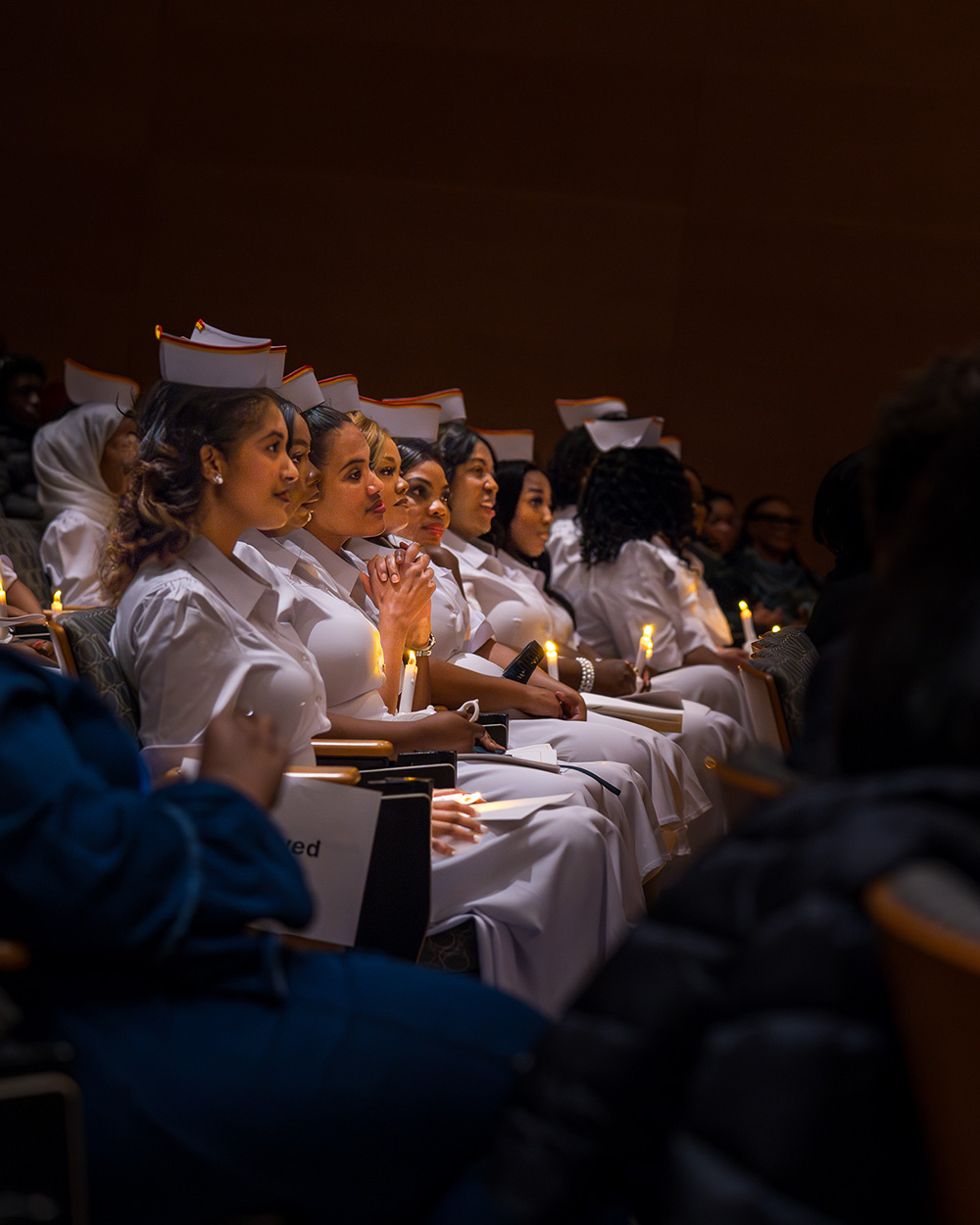 Nursing graduates wearing white nurses uniforms and holding candles in a darkly lit auditorium.
