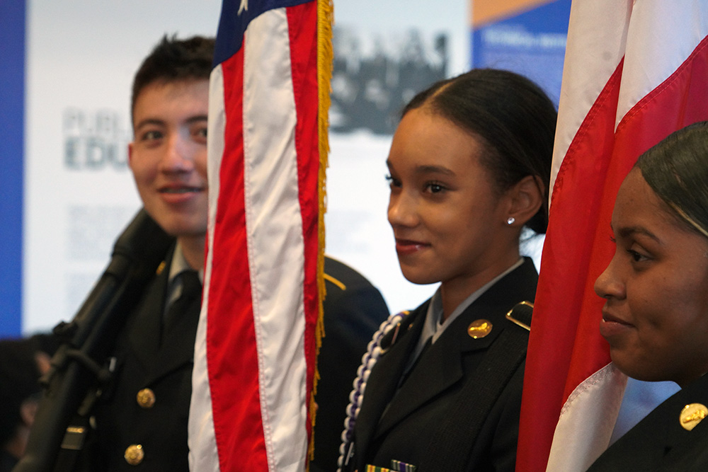 Three students in military attire holding US flags and smiling.