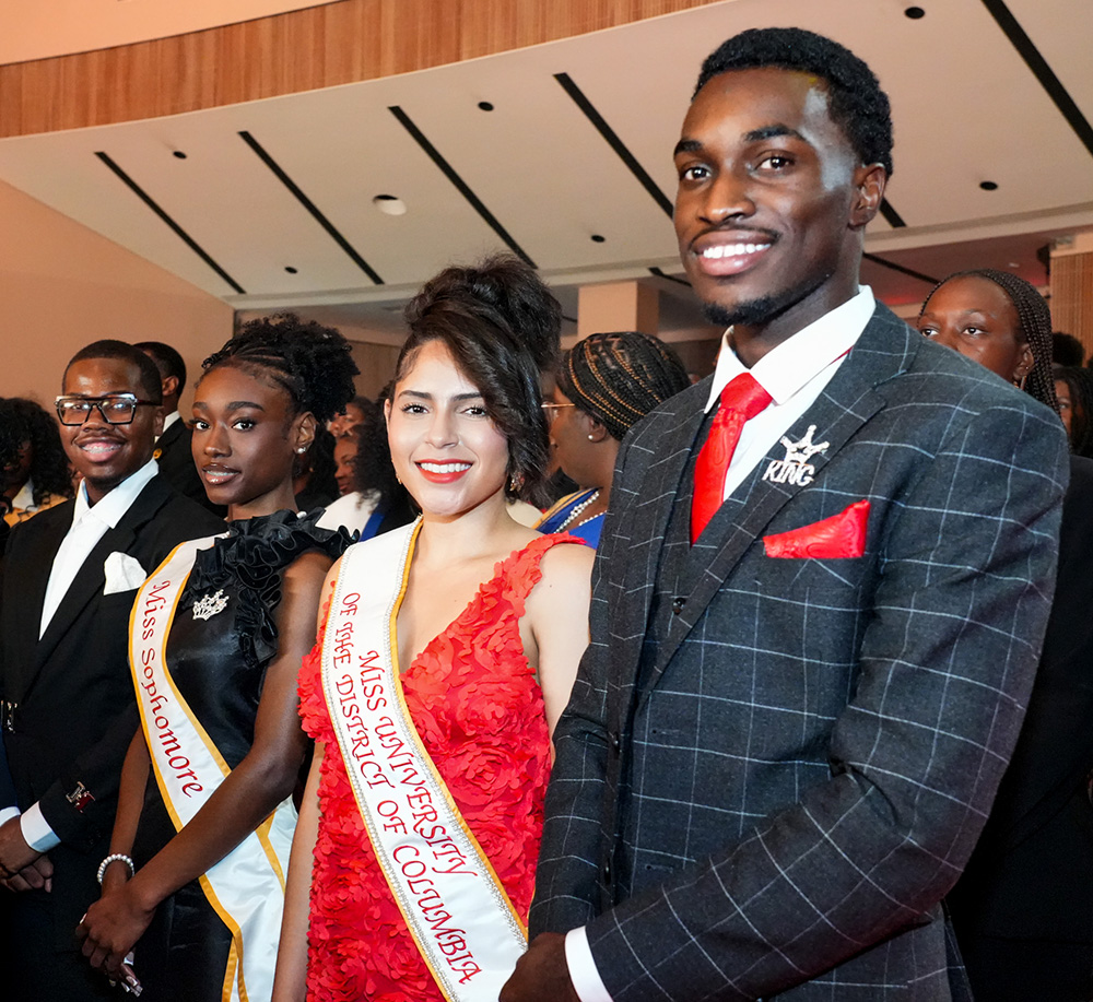 Students dressed in formal attire at a campus pageant, including two wearing sashes reading “Miss Sophomore” and “Miss University of the District of Columbia.”