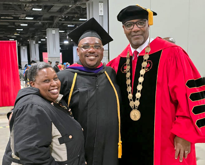 Three people pose at a graduation ceremony: a graduate in cap and gown stands between a smiling companion and a university official in red academic regalia with a ceremonial chain.
