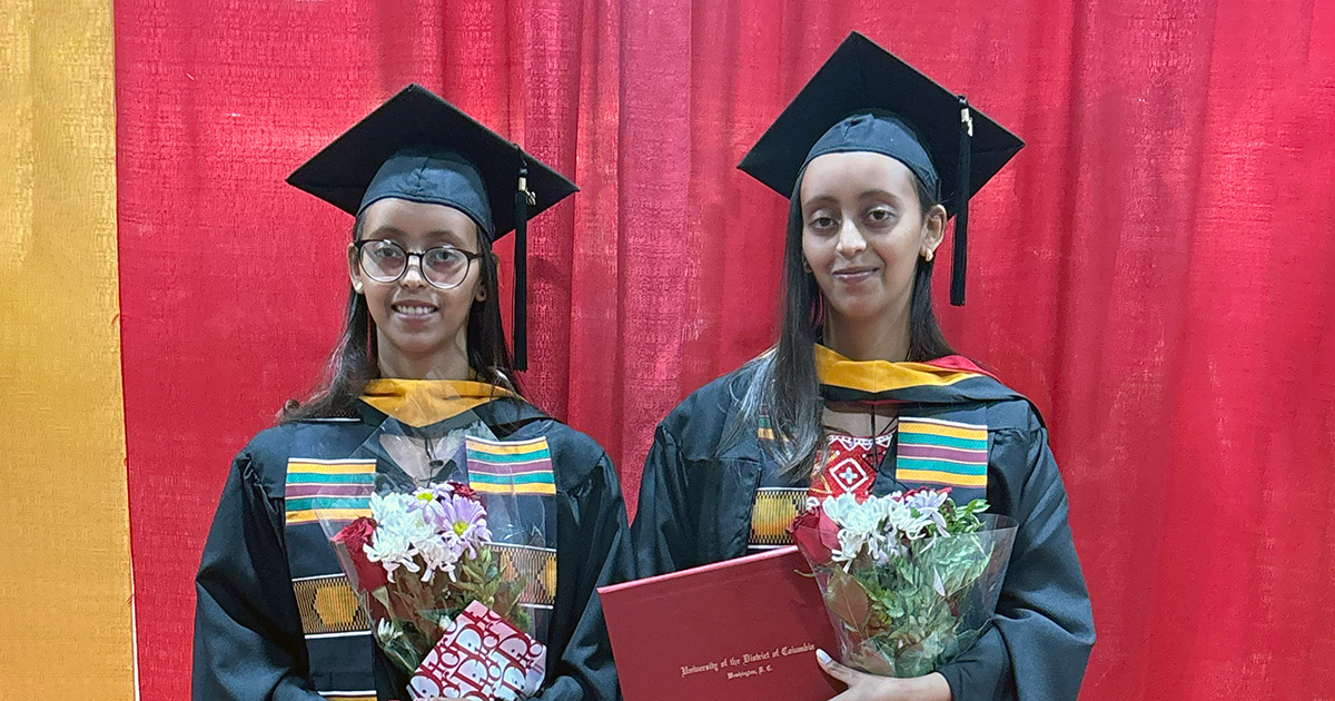 Twin females standing in graduation cap and gown in front of a red and yellow backdrop.