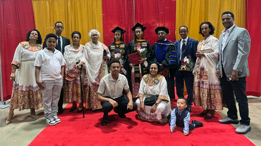 Large family group portrait on a red carpet in front of red and gold curtains, with several people in graduation caps and gowns holding flowers.