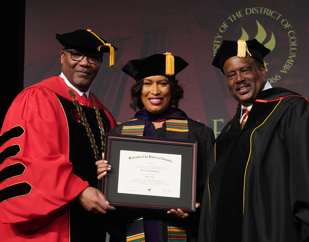 Three people dressed in graduation regalia standing together and smiling. One holds a framed certificate.