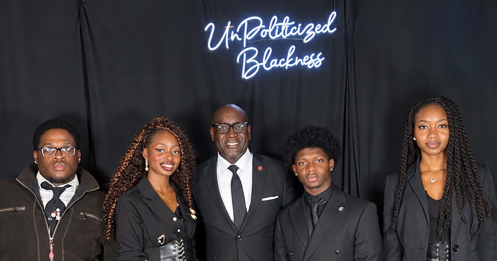Five people dressed in black formalwear pose in front of a black backdrop with a neon sign reading “UnPoliticized Blackness.”