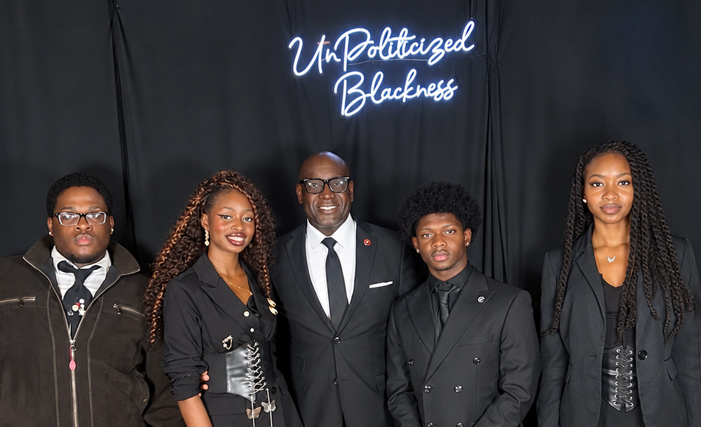 Five people dressed in black formalwear pose in front of a black backdrop with a neon sign reading “UnPoliticized Blackness.”