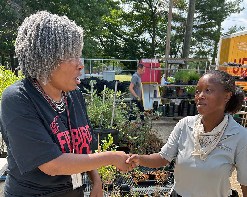 Two people shake hands while talking at an outdoor plant nursery, with potted plants and shelves in the background.