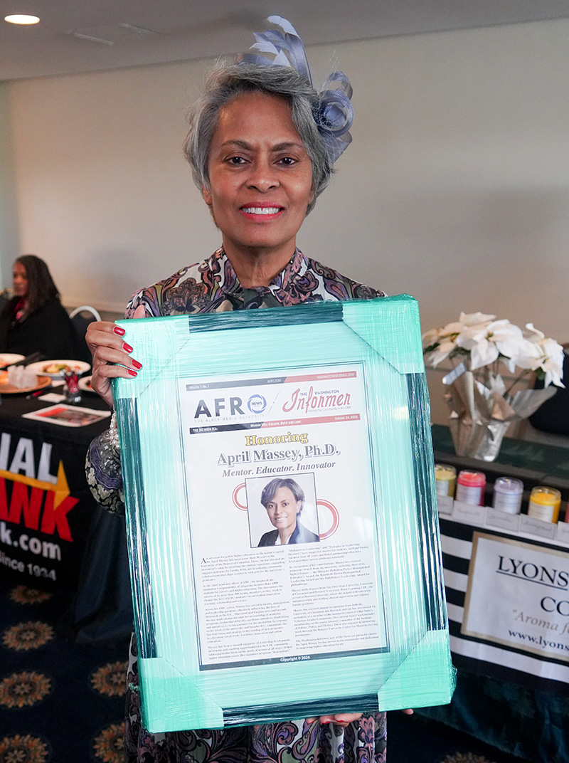 Woman wearing a decorative hat holds a large framed newspaper tribute featuring her portrait at an indoor event.