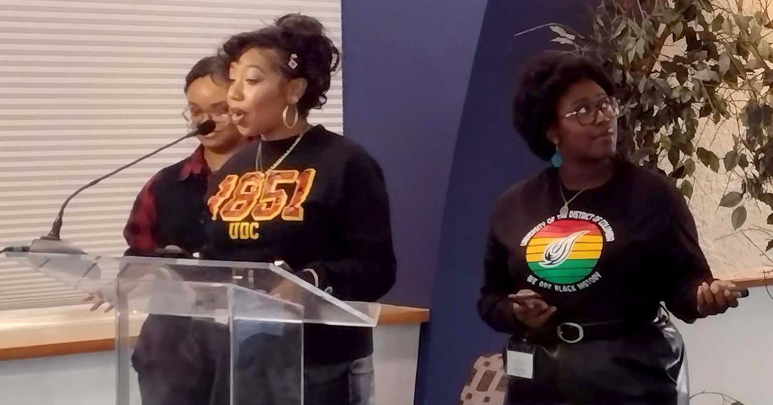 Three people at a podium during an indoor event, with one person speaking into a microphone while another stands to the side holding a phone.